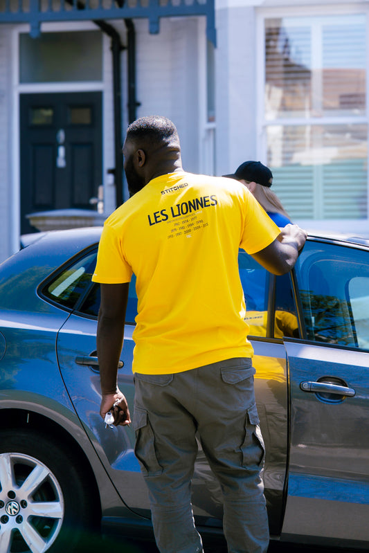Man wearing a bright yellow t-shirt with STITCHED at the collar and “LES LIONNES” printed above a list of years on the back, opening the rear door of a gray sedan parked in front of a house.