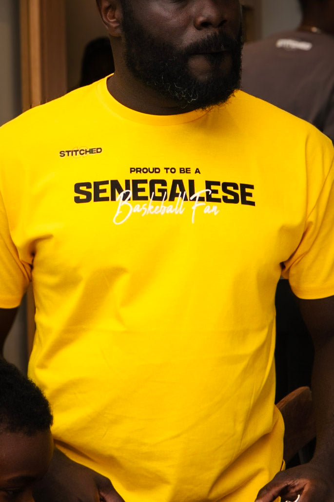 Man wearing a bright yellow t-shirt with STITCHED on the upper left and “Proud to be a Senegalese Basketball Fan” printed across the chest.