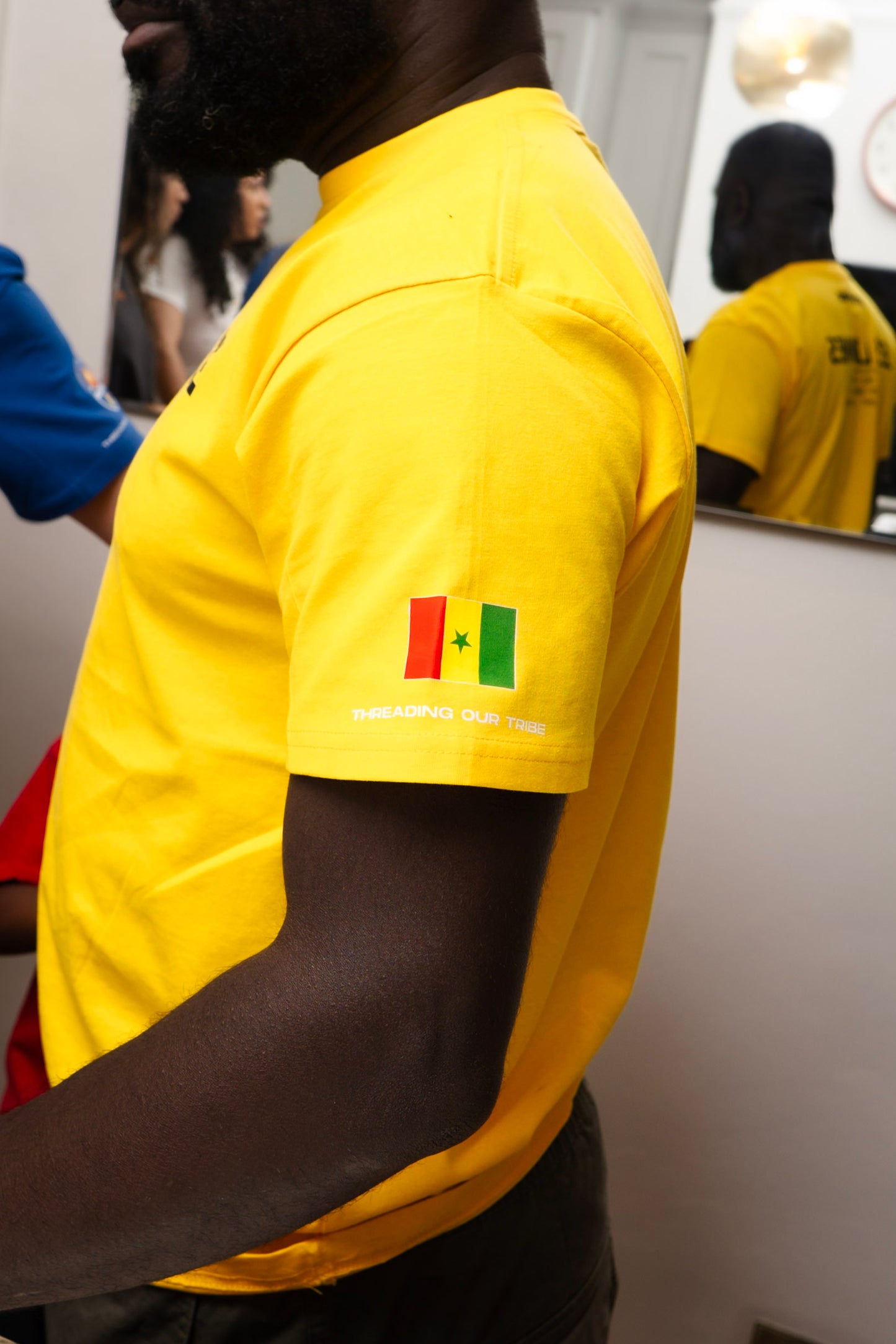 Man wearing a bright yellow STITCHED t-shirt with a small Senegalese flag graphic and “Threading Our Tribe” printed on the sleeve, standing indoors with his reflection visible in a wall mirror.