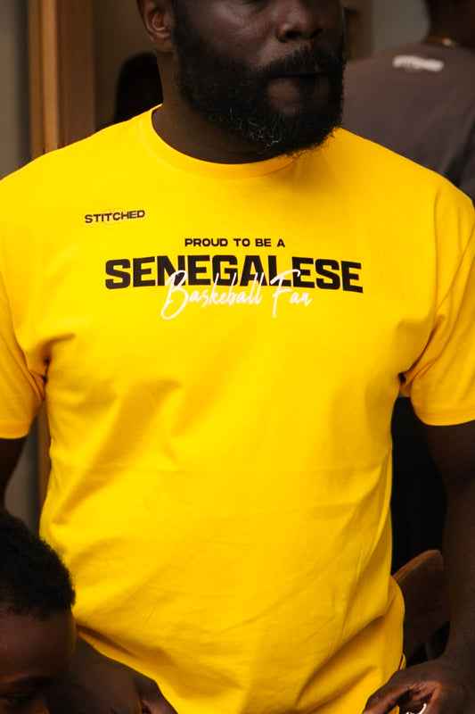 Man wearing a bright yellow t-shirt with STITCHED on the upper left and “Proud to be a Senegalese Basketball Fan” printed across the chest.