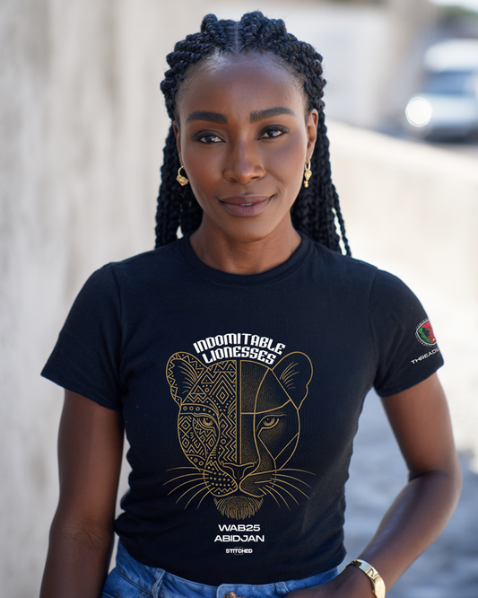 Female model wearing The Mask Tee in black, featuring a half-mask, half-court-line lioness graphic with “Indomitable Lionesses” header and “WAB25 Abidjan” branding, posed outdoors.